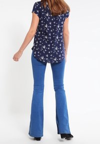 Tmavě modrá blůza s krátkým rukávem s ptačím vzorem, v kombinaci s modrými rozšířenými džíny. Oblečení má uvolněný střih a hladkou strukturu.