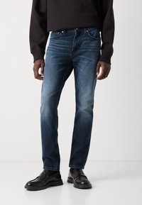 Jeans de mezclilla azul oscuro con corte recto y acentos desgastados. Cuenta con cinco bolsillos y cierre con botón, llevados con mocasines negros.