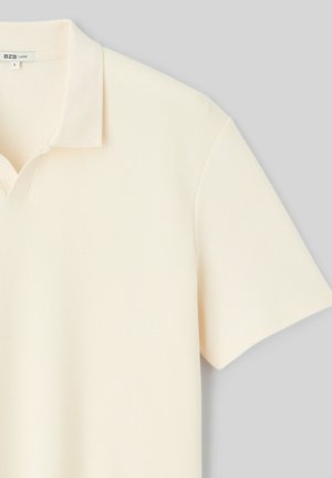 Polo à manches courtes côtelé de couleur crème avec col et coupe ample, étiquette taille grande, sur fond gris clair.