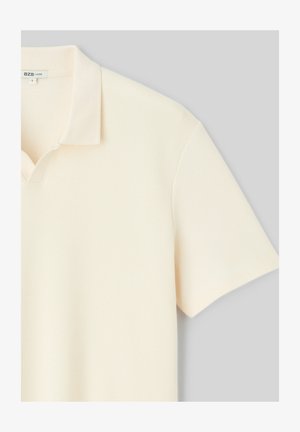 Polo à manches courtes côtelé de couleur crème avec col et coupe ample, étiquette taille grande, sur fond gris clair.