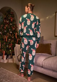 Completo pigiama verde con motivo ripetuto di volti di Babbo Natale con cappelli rossi e bianchi. Tessuto morbido, vestibilità ampia e polsini elastici.