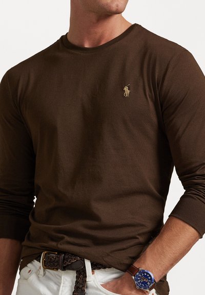 Brown long-sleeve shirt made of cotton, featuring a small gold logo on the chest. Paired with a braided belt and a blue watch.