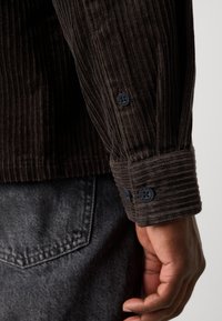 Dark brown corduroy shirt with a ribbed texture, featuring black buttons on the cuffs. The shirt is paired with gray denim pants.