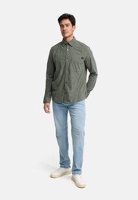 Camisa de pana verde con botones y bolsillo, combinada con jeans azul claro y zapatillas blancas. La camisa tiene una superficie texturizada.