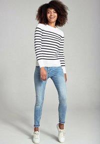 Maglione a righe con maniche lunghe, a righe navy e bianche, abbinato a jeans skinny azzurri e sneakers bianche, in piedi su uno sfondo chiaro.