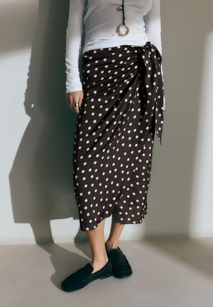 Mujer con una blusa blanca de manga larga y una falda midi negra con lunares blancos, combinada con zapatos negros sin cordones y joyas doradas.