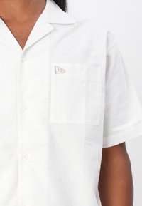 Camicia bianca a maniche corte con un design a bottoni, tasca sul petto sinistro e logo ricamato in beige tenue. Tessuto morbido.