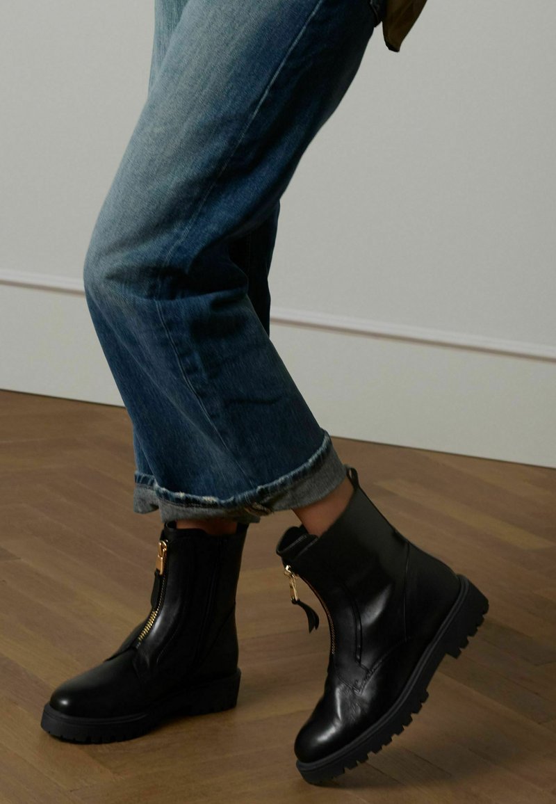 Stivaletti neri in pelle con suola spessa e dettaglio della cerniera dorata. Indossati con jeans blu arrotolati, che mettono in risalto la texture e la lucentezza degli stivali.