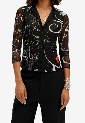 Chemise noire transparente à boutons avec un dessin abstrait de visage en lignes blanches et de petites touches de couleur, portée avec un pantalon noir et des bijoux en argent.
