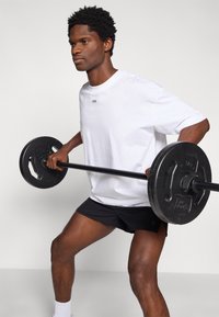 Λευκός κοντομάνικος πουκάμισο με μικρό λογότυπο στο στήθος, συνδυασμένο με μαύρο σορτς, σηκώνοντας μια μαύρη μπάρα με κυκλικά βάρη.