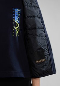 Navy ribbad t-shirt med en texturerad puffärm; har färgglad branding. Puffärmen är slät och vadderad med en logotyp på ärmslutet.