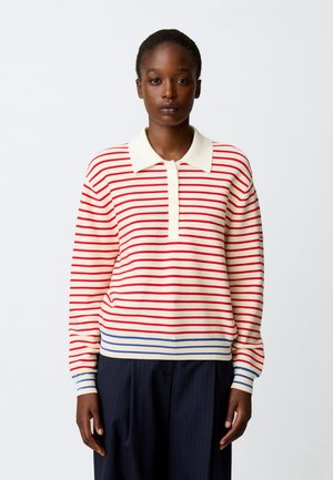 Donna che indossa un maglione crema a maniche lunghe con strisce orizzontali rosse, un colletto bianco, strisce blu sull'orlo e sui polsini, e pantaloni scuri.