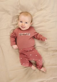 Otroška obleka rožnate barve z dolgimi rokavi, z napisom "SISTER" v cvetličnem potisku, v kombinaciji s hlačami z pikami. Mehka tkanina, položena na bež ozadje.