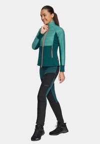 Zielona i ciemnozielona kurtka zapinana na zamek, z pikowaną przednią częścią, w połączeniu z czarnymi spodniami z boku z zamkami błyskawicznymi i akcentami logo. Uzupełniona czarnymi botkami.