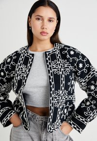Čierna látková bunda s bielou kvetinovou výšivkou, okrúhlym výstrihom a ľahkým dizajnom. Modelka má pod ňou oblečený šedý krátky top.