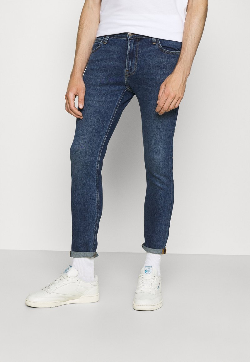 Slim-fit blue denim jeans with a subtle wash, five-pocket design, and rolled cuffs at the ankles, paired with white sneakers.