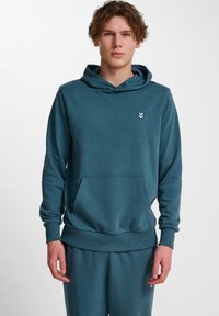 Felpa in cotone color teal con cappuccio regolabile, tasca a marsupio, polsini e orlo a coste. Presenta un piccolo logo a forma di freccia bianca sul petto.