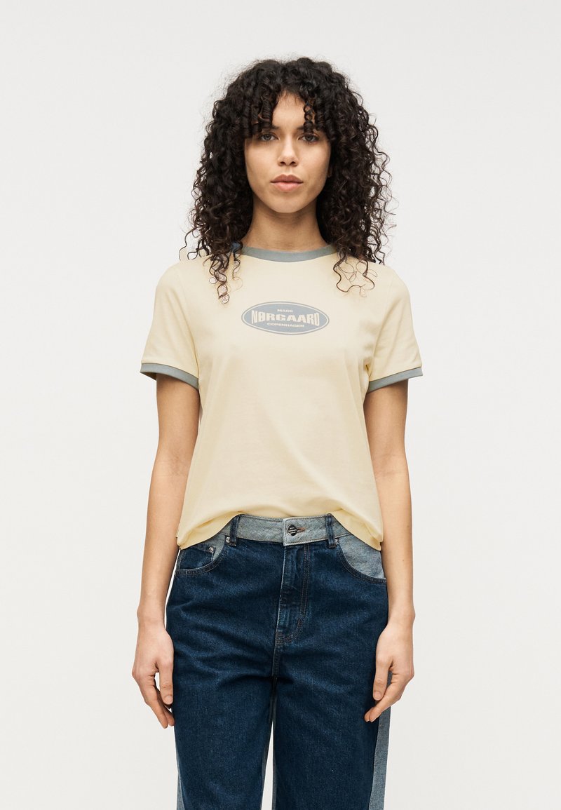 Ανοιχτό κίτρινο μπλουζάκι με γκρι ριμπ στη λαιμόκοψη και τα μανίκια, που φέρει ένα κεντρικό λογότυπο σε σκούρο γκρι. Φορεμένο με σκουρό μπλε τζιν.