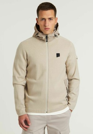 Beige Zip-Jacke mit Kapuze aus strukturiertem Stoff. Verfügt über ein schwarzes Logo auf der Brust und zwei seitliche Taschen. Einfaches, funktionales Design.