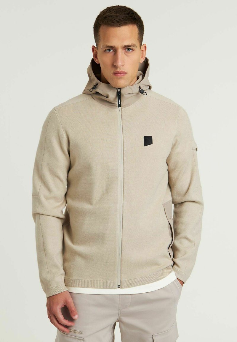 Chaqueta beige con cremallera y capucha, hecha de tela texturizada. Presenta un logo negro en el pecho y dos bolsillos laterales. Diseño simple y funcional.