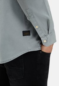 Hellgraues, knöpfbares Hemd aus glattem Stoff mit schmaler Passform, gesteppten Details und einem schwarzen Etikett mit goldfarbener Schrift an der Seite.