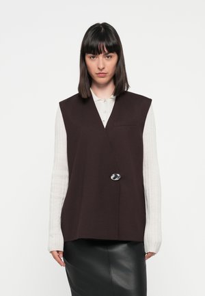 Dark brown sleeveless vest with a single metallic button, paired with a light ribbed long-sleeve shirt, completing a sleek silhouette.