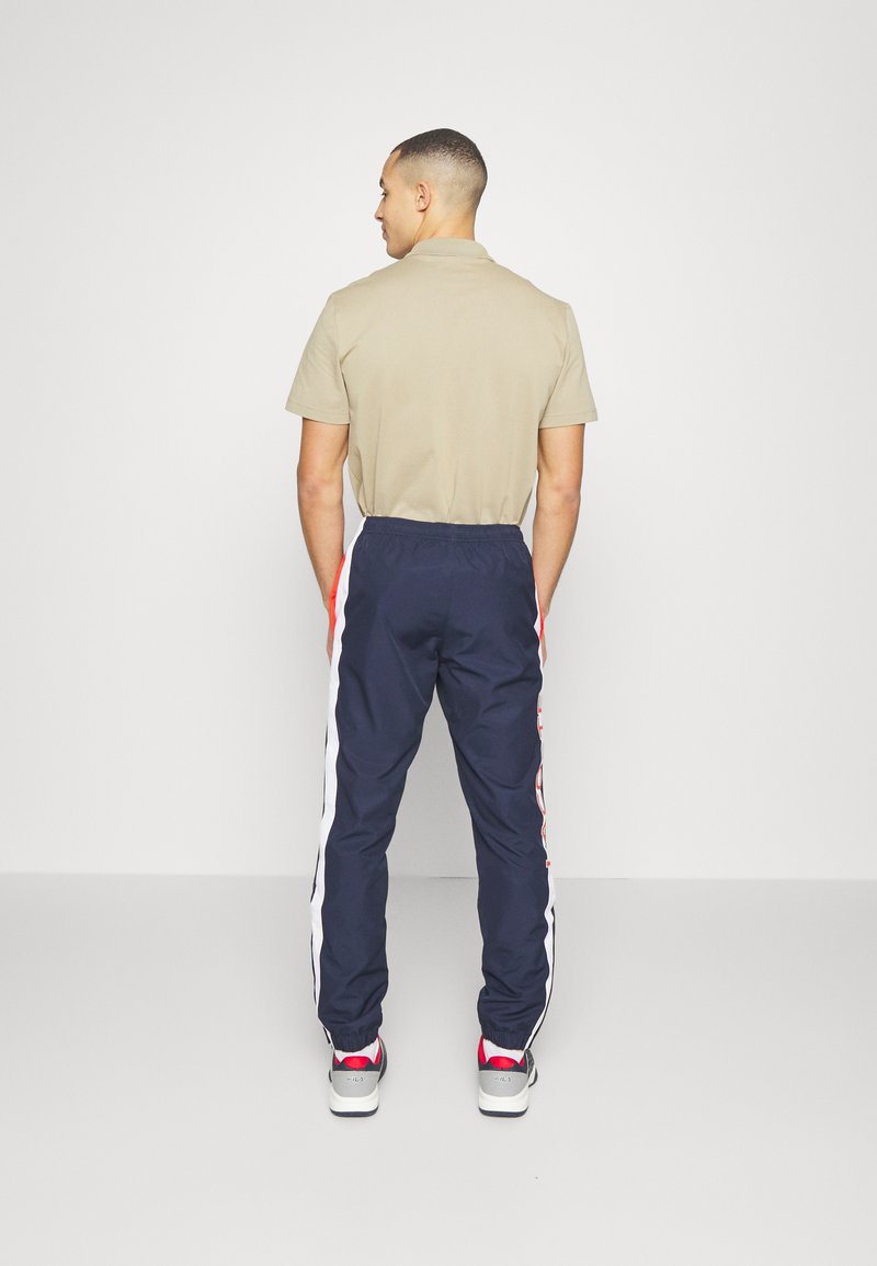 Homme de dos portant un polo beige, un pantalon de survêtement bleu marine avec des rayures blanches et rouges sur le côté, et des baskets blanches et rouges sur fond uni.