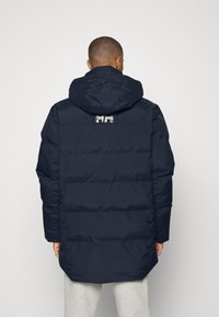 Jachetă puffer de culoare bleumarin, cu glugă, mâneci lungi și design matlasat orizontal. Detaliu cu logo vizibil pe partea superioară a spatelui. Textură netedă.