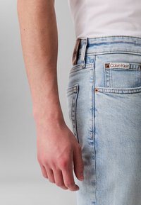 Gaiši zili džinsa džinsi ar taisnu piegriezumu, ar vara detaļām un brūnu ādas uzlīmi ar uzrakstu "Calvin Klein" jostasvietā.