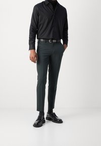 Zwarte langemouwige overhemd met knopen, donkergroene op maat gemaakte broek, zwarte leren riem en zwarte veterschoenen met dikke zolen.