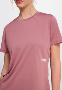 Jasnoróżowa, krótkoszarlotkowa koszulka sportowa z prążkowaną fakturą oraz białym logotypem po lewej stronie, posiadająca dekolt typu crew neck.