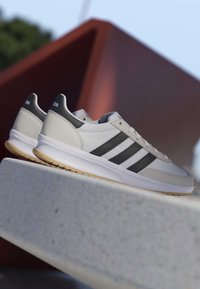 Weiße Sneaker mit schwarzen Streifen und Gummisohlen, die auf einer Betonkante vor einem verschwommenen Außenhintergrund platziert sind.