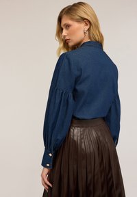 Camicia di denim scuro con maniche a sbuffo e polsini bottone, abbinata a una gonna a pieghe in pelle marrone. Texture liscia e design strutturato.