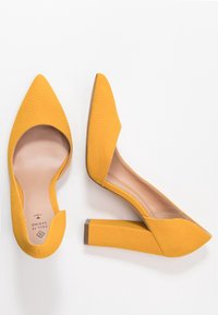 Escarpins jaunes à talons hauts avec une finition texturée, un bout pointu, des côtés ouverts et un talon épais, dotés d'une doublure intérieure lisse.