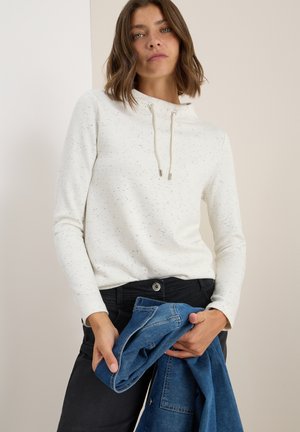 Sweatshirt blanc avec texture tachetée, col montant et cordons de serrage. Assorti avec un jean noir, tenant une veste en denim bleu.