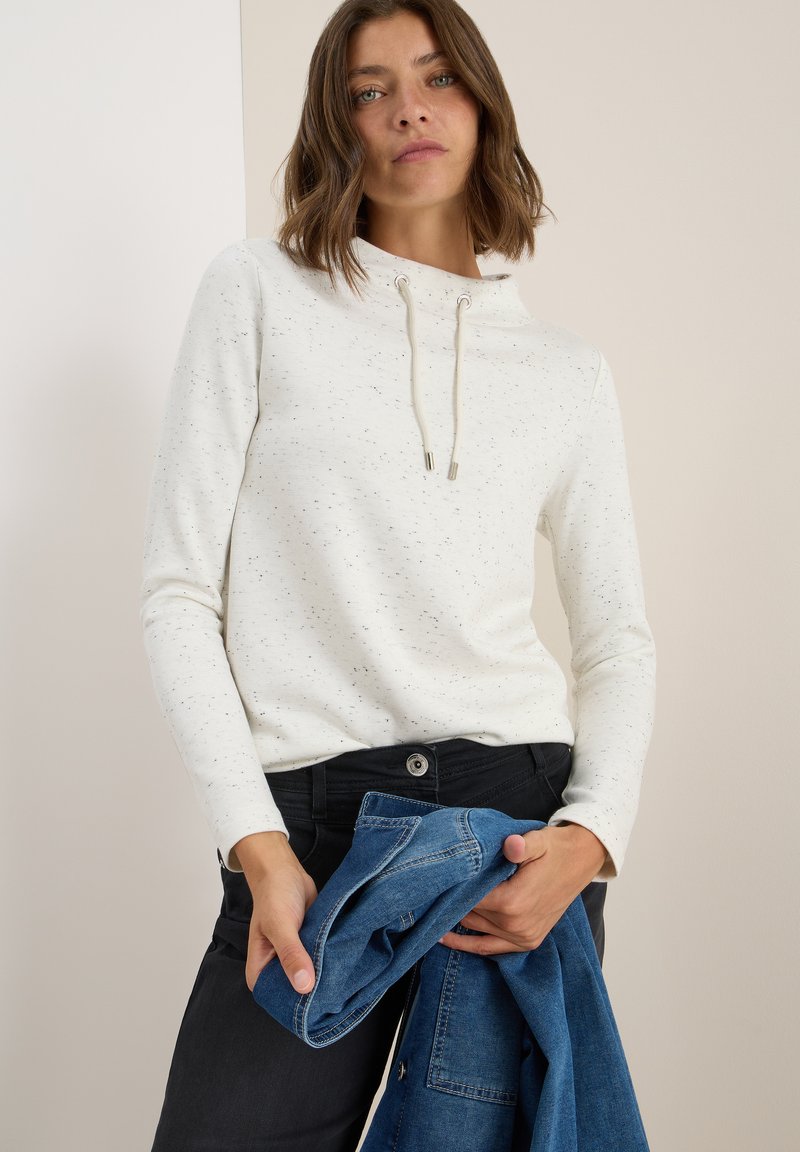 Vit sweatshirt med prickig textur, hög krage och dragsnören. Matchas med mörka jeans och håller en blå denimjacka.