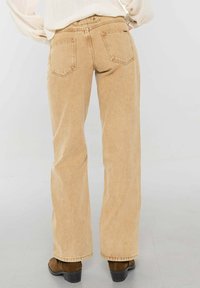 Jean beige taille haute à jambes larges en denim, avec deux poches arrière, coutures discrètes et texture lisse. Associé à des bottines marron.