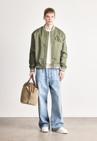 Olivrohelise bomber-tüki jope triibulise kraega, valge kootud sviiter, helesinised laiapõlvalised teksad ja pruun duffelkott. Valged tossud.