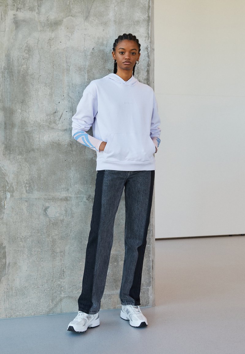 Felpa viola chiaro con tasca frontale, accenti blu sulle maniche; abbinata a jeans grigi scuri. Sneakers bianche completano il look.