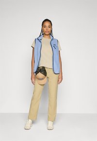 Kvinne med flettet hår, iført en lyseblå fleecevest, beige t-skjorte, khaki bukser og hvite joggesko, holder en beige og svart caps ved siden av.
