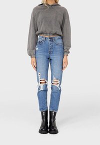 Sudadera corta gris con dobladillo elástico, combinada con vaqueros azules desgastados que tienen múltiples desgastes y botas negras hasta el tobillo con detalles de cremallera.