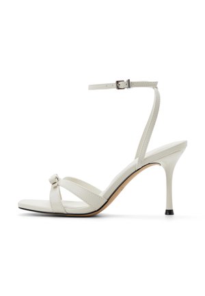 Sandali bianchi con cinturini, tacco alto con dettaglio a nodo, cinturino alla caviglia regolabile, texture liscia in pelle e tacco con punta nera.