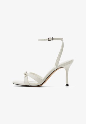 Sandalias de tacón alto blancas con tiras, detalle de nudo, correa ajustable en el tobillo, textura de cuero suave y tacón rematado en negro.