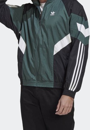 Mężczyzna noszący zieloną, czarną i białą kurtkę Adidas na suwak z logo na piersi i trzema paskami na rękawach, w połączeniu z czarnymi spodniami.