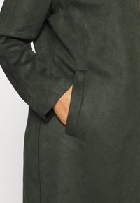 Grön mockajacka med en slät textur, med en sidoficka och långa ärmar, som framhäver en ren, minimalistisk design.