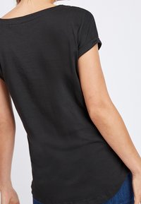 Kvinde iført en simpel sort kortærmet T-shirt med opkrøllede ærmer, set fra ryggen, kombineret med blå jeans foran en hvid baggrund.