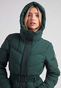 Chaqueta acolchada verde con un cuello alto y capucha. Presenta costuras horizontales, una cintura con cinturón y un parche de logo en el hombro.