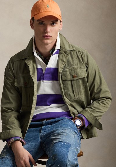 Olívazöld dzseki fehér és lila csíkos póló fölött, kék, kopott hatású farmernadrág, narancssárga sapka. Látható óra és kockás részlet.