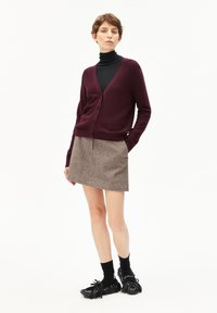 Vrouw staat met een zwarte coltrui, bordeauxrode cardigan, grijze rok, zwarte sokken en zwarte mocassins met franjes tegen een witte achtergrond.