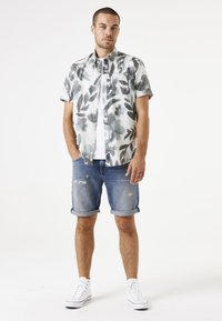 Korte mouwen overhemd met een bloemmotief in grijs en groen op een witte achtergrond, gecombineerd met opgerolde denim shorts en witte sneakers.